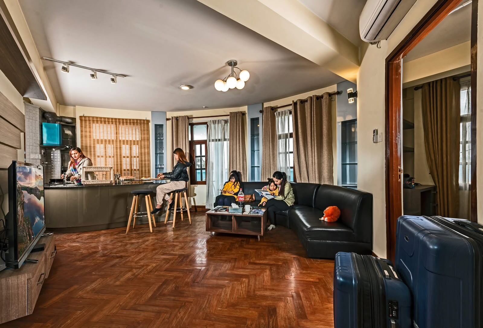 Guests in an apartment living hall: an open kitchen with a breakfast bar, a corner sofa, and suitcases by the door.